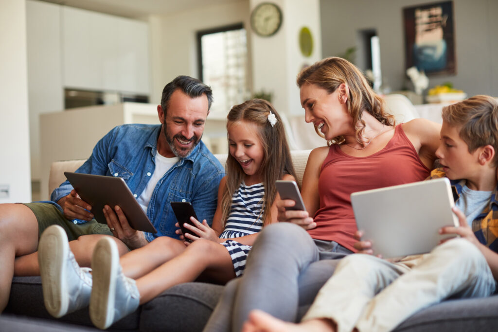 En glad familj beståendes av en man, kvinna och två barn som sitter i en soffa med varsin digital enhet.