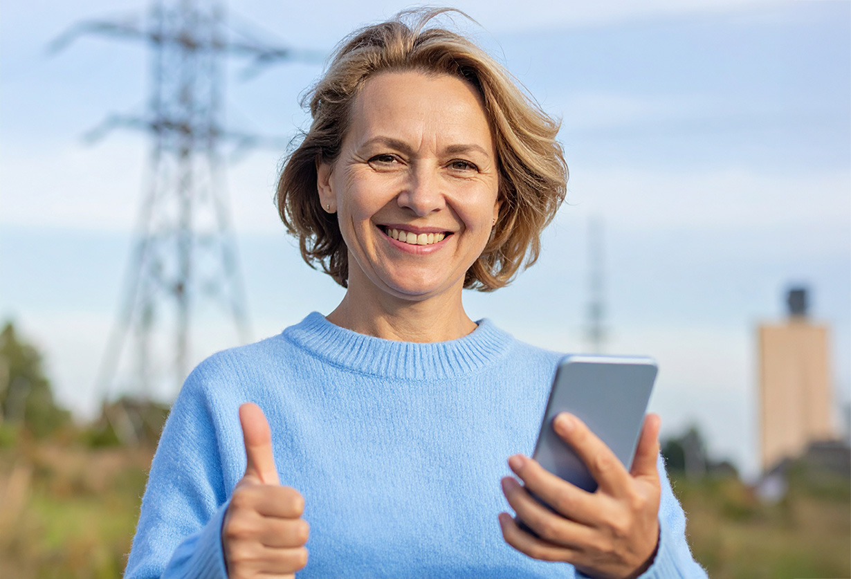 En kvinna håller en mobiltelefon i handen och gör tummen upp med en elledning i bakgrunden.