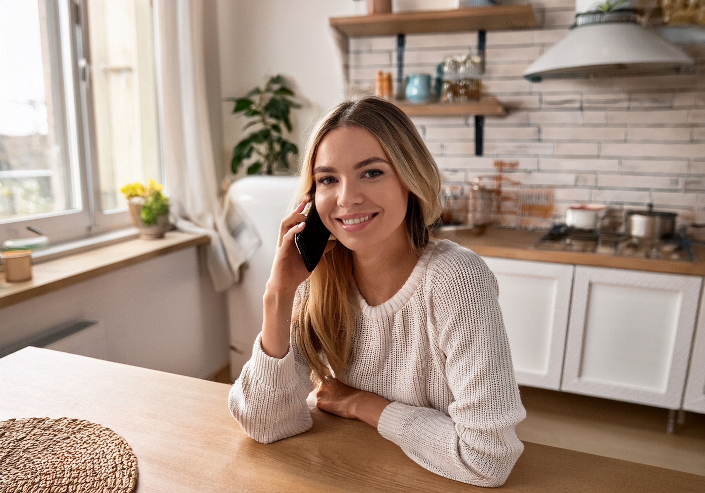 Kvinna sitter vid köksbordet och pratar i telefon och ser glad ut