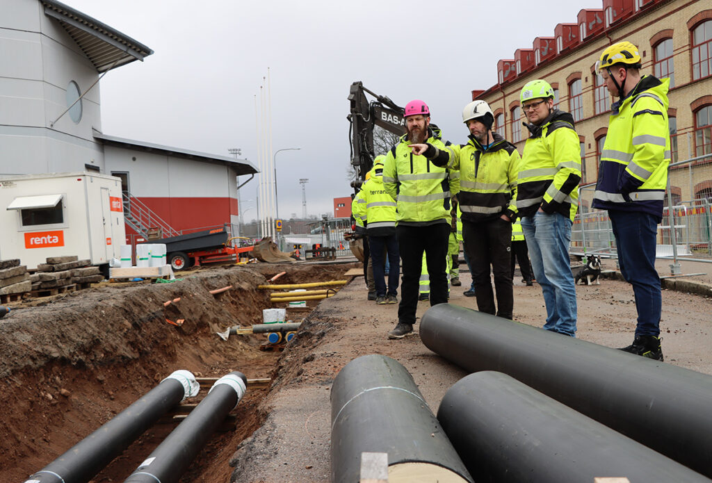 Mattias Sandström och Mariia Zetterberg står intill schaktet med projektgrupppen och grävmaskinen i bakgrunden.
